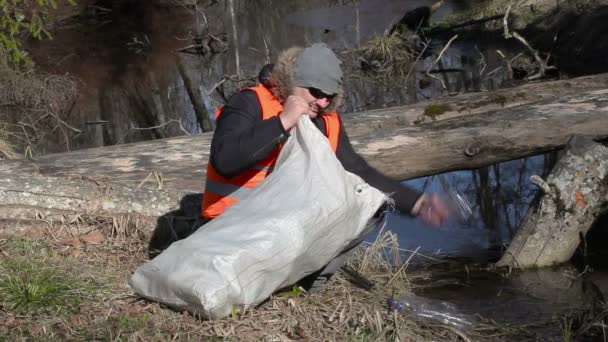 Чоловік з сумкою збирає порожні пластикові пляшки біля річки — стокове відео