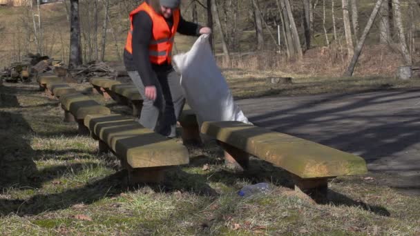 Чоловік з сумкою збирає порожні пластикові пляшки біля лавок в парку — стокове відео