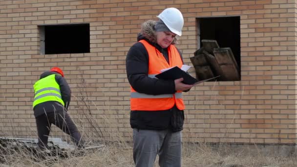 Gerente de obra con documentación y trabajador en segundo plano cerca del edificio — Vídeo de stock