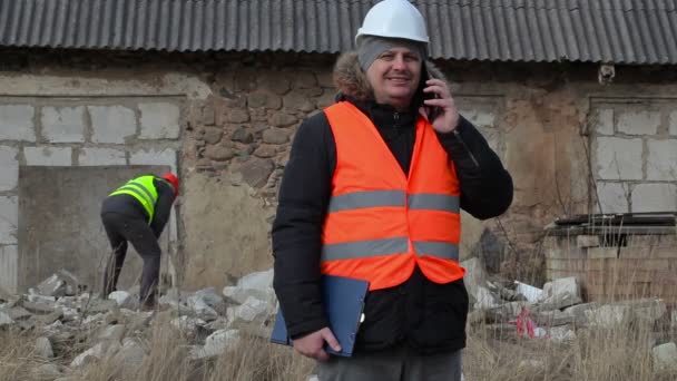 Ispettore di costruzione parlando su smartphone in background lavoratore utilizzando mazza — Video Stock