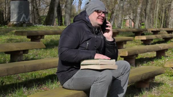 Homem com dois livros e telefone inteligente no banco no parque — Vídeo de Stock