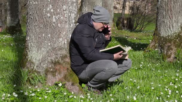 Człowiek, czytania książki i rozmowy na inteligentnego telefonu w łące w pobliżu drzewa — Wideo stockowe