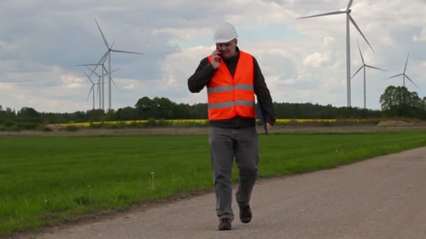 Yürüyüş ve yel değirmenleri yakınındaki akıllı telefonda konuşurken elektrikçi mühendisi — Stok video