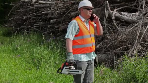 Лесоруб с бензопилой разговаривает на смартфоне возле кучи ветвей — стоковое видео