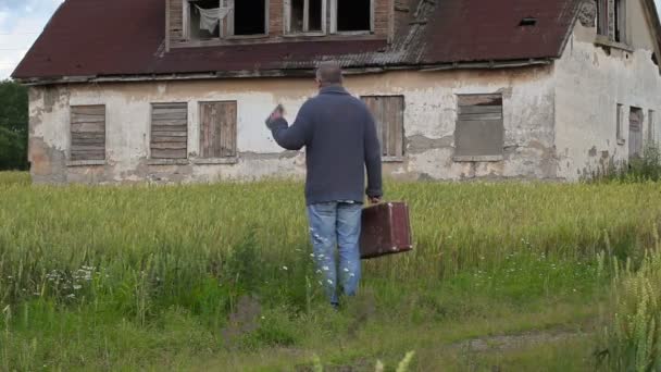 Человек с чемоданом смотрит на старый заброшенный дом и уходит — стоковое видео