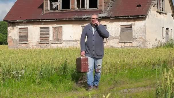 Мужчина с чемоданом разговаривает по смартфону возле заброшенного здания — стоковое видео