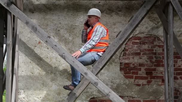 Travailleur de la construction assis sur un échafaudage en bois et parlant au téléphone — Video
