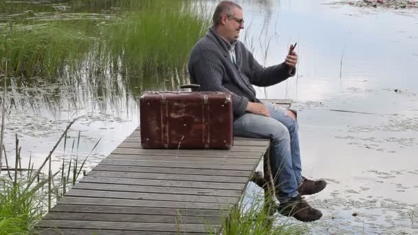 Mannen med resväska sitter nära lake och pratar på telefon — Stockvideo