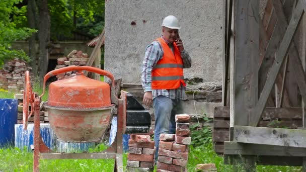 Trabajador de la construcción hablando por teléfono cerca de andamios de madera y hormigonera — Vídeo de stock