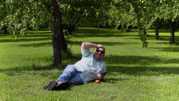 Man with tablet PC laughing and lying at apple orchard — Stock Video