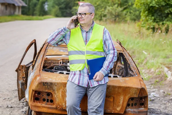 Inspector hablando por teléfono inteligente cerca quemado restos de coche en el lado de la carretera —  Fotos de Stock