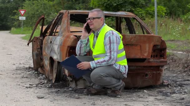 Инспектор говорит по смартфону около сгоревшей автокатастрофы на обочине — стоковое видео