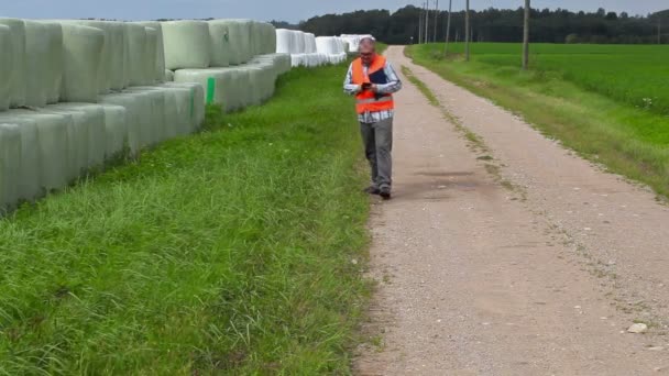 Фермер с помощью планшетного ПК и ходить возле тюков сена — стоковое видео