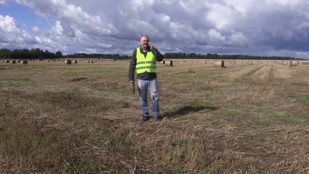 Agricultor hablando en el teléfono inteligente en el campo cerca de fardos de paja — Vídeos de Stock