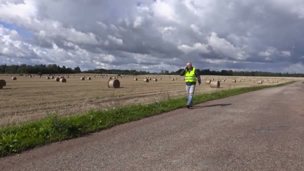 Agricultor con smartphone caminando por la carretera cerca de fardos de paja — Vídeo de stock