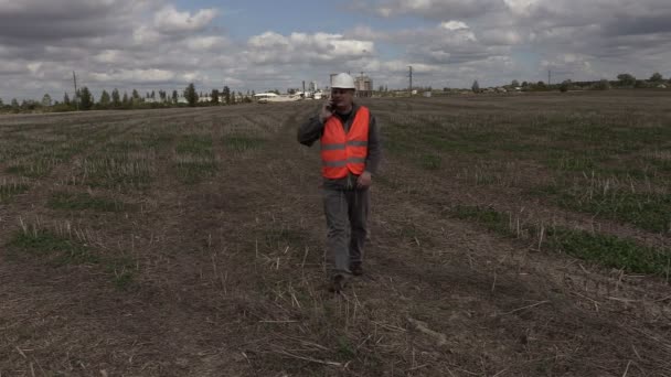 Ingenieur praten op de smartphone en lopen op het veld in de buurt van fabriek — Stockvideo