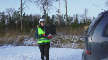 Kadın orman bilim adamı dizüstü bilgisayar tutuyor ve arabanın yanında yürüyor.