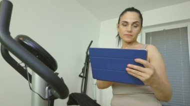 Evde tablet kullanan ve egzersiz bisikleti kullanan bir kadın.