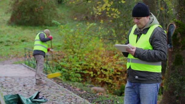 Förman arbetar med tablet Pc nära arbetstagare som samla löv — Stockvideo