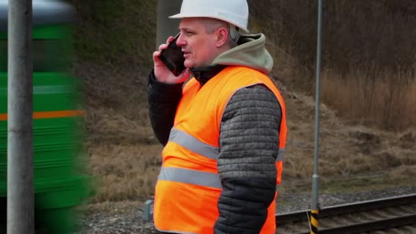 Сотрудник железной дороги разговаривает по мобильному телефону возле проходящего поезда — стоковое видео