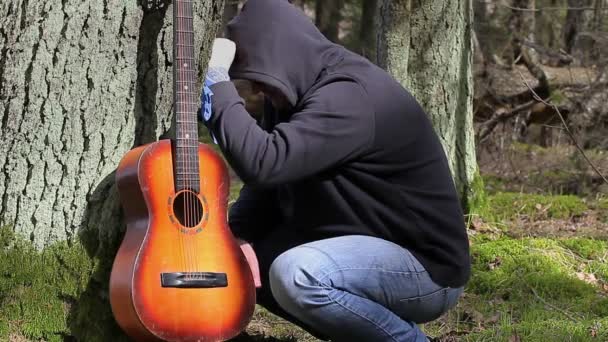 Стрессоустойчивый мужчина с гитарой в лесу возле дерева — стоковое видео