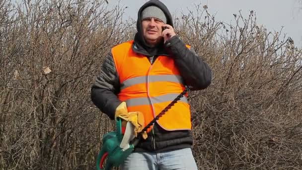 Liggande arbetare med bush cutter talar i mobiltelefon — Stockvideo