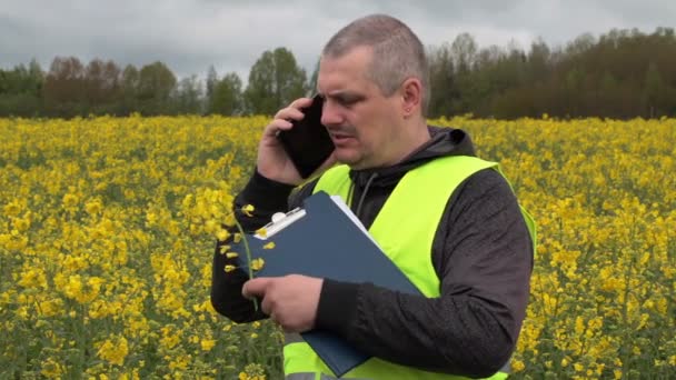 Rolnik z telefonem komórkowym w pobliżu żółtego rzepaku pola — Wideo stockowe