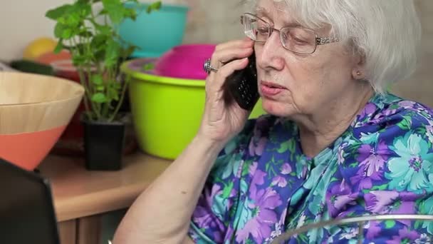 Señora mayor hablando en el teléfono celular cerca del ordenador portátil — Vídeos de Stock