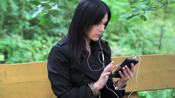 Женщина слушает музыку в парке на скамейке — стоковое видео