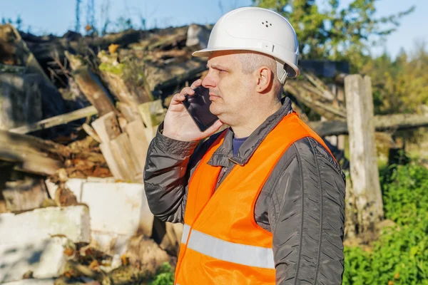 Inspector de construcción hablando en el teléfono inteligente cerca de abandonado, edificio dañado —  Fotos de Stock