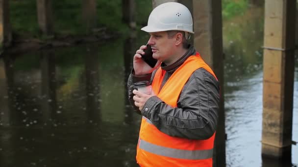 Рабочий пьет кофе и разговаривает на смартфоне под мостом — стоковое видео