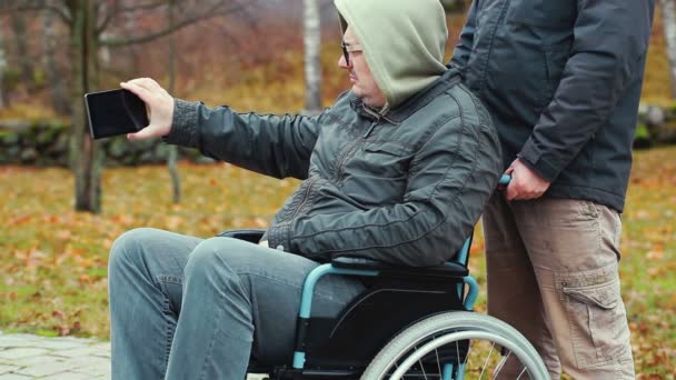 Homme handicapé filmé avec smartphone en fauteuil roulant avec assistant derrière — Video