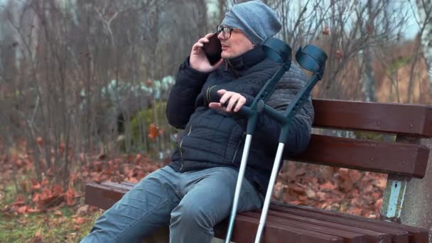 Homem com deficiência com muletas e smartphone no banco — Vídeo de Stock