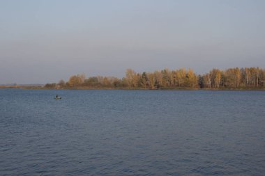 Sakin bir nehir ve teknede yalnız bir balıkçı olan sonbahar manzarası. Arka planda renkli bir orman.