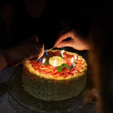 Doğum günü pastası ve yanan mumlar. Karanlık odada doğum günü pastasında mum yakan iki el.