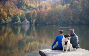 Sonbahar gölünün kenarında oturan iki adam ve köpek.