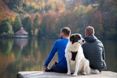Sonbahar gölünün kenarında oturan iki adam ve köpek.