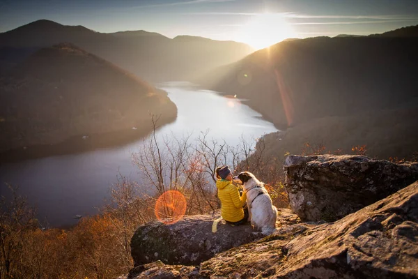 夕暮れ時には湖の景色を眺めながら岩の上に座っている女性と犬 — ストック写真