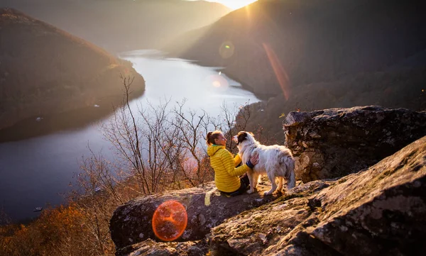 夕暮れ時には湖の景色を眺めながら岩の上に座っている女性と犬 — ストック写真