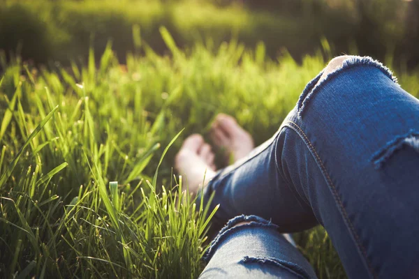 穿着破牛仔裤的嬉皮士女人的腿 躺在绿草中 戴着智能手机和耳机 享受夕阳西下 夏天的放松 — 图库照片