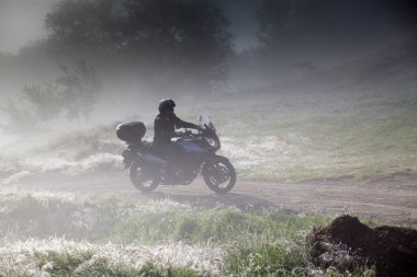 Sabahın erken saatlerinde sisli yolda motosiklet süren bir adamın silüeti.