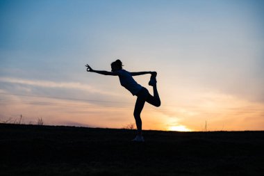 Gün batımında denge pozisyonunda olan bir kadın dışarıda yoga yapıyor.