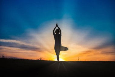 Gün batımında denge pozisyonunda olan bir kadın dışarıda yoga yapıyor.