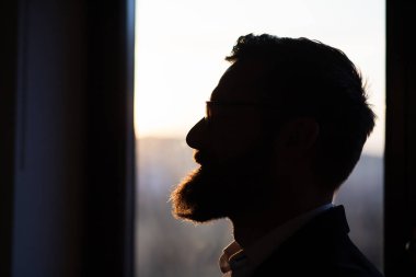 Sakallı adam pencerede gün batımına karşı duruyor.  