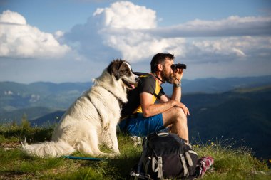 Yakışıklı adam ve beyaz köpek, dürbünle doğada ağır ağır ilerliyorlar.