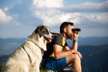 Yakışıklı adam ve beyaz köpek, dürbünle doğada ağır ağır ilerliyorlar.