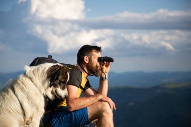 Yakışıklı adam ve beyaz köpek, dürbünle doğada ağır ağır ilerliyorlar.
