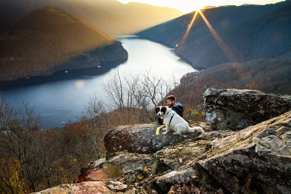 夕暮れ時には湖の景色を眺めながら岩の上に座っている男と犬 — ストック写真