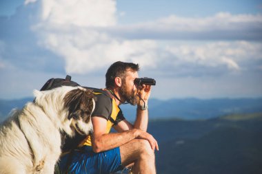 Yakışıklı adam ve beyaz köpek, dürbünle doğada ağır ağır ilerliyorlar.