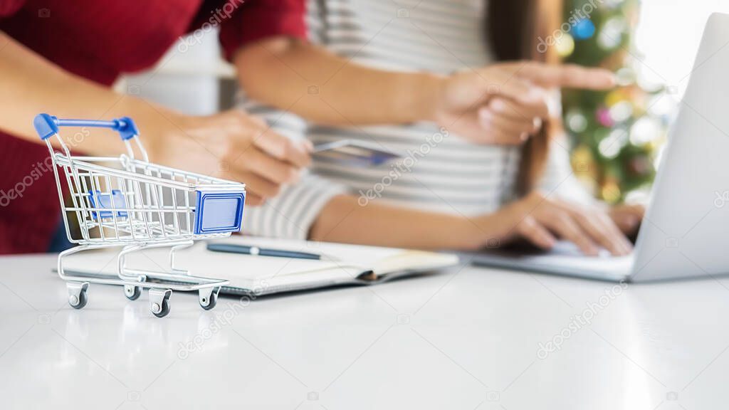 Online shopping concept. Shopping cart on white table with Asian woman holding credit card and using tablet for online shopping and Christmas tree background.